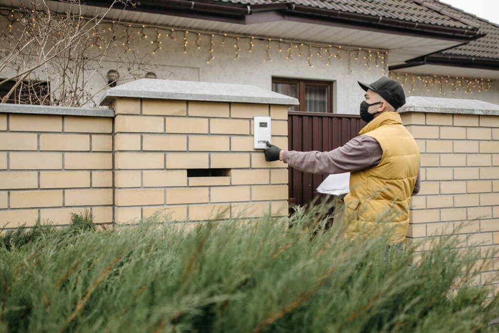 A delivery worker wearing a face mask presses a doorbell at a residential home, emphasizing contactless service.